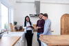 a man and his male partner in a kitchen look at a tablet that a business woman is showing them